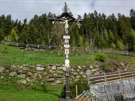 Ein kunstvoller hölzerner Kreuzweg mit verschiedenen Figuren und Symbolen. Im Hintergrund erstreckt sich eine grüne Wiese und bewaldete Hügel. | © Bernhard Gruber – Photography