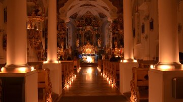 Eine stimmungsvolle Kirche mit gedecktem Licht und verzierten Säulen. Der Altar ist im Hintergrund sichtbar und von Kerzen beleuchtet.