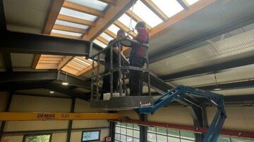 Zwei Arbeiter stehen in einer Hebebühne und reparieren das Dach eines großen Industriegebäudes. Im Hintergrund sind Werkstattmaschinen und große Fenster zu sehen.