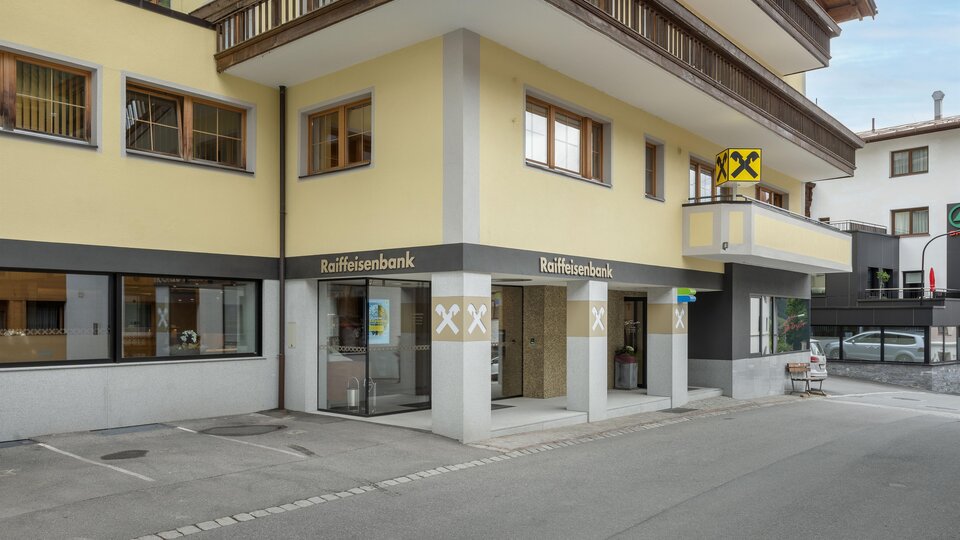 Ein modernes Bankgebäude mit gelber Fassade und großen Fenstern. Im Vordergrund ist ein Parkplatz und eine Straße sichtbar.