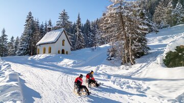 Zwei Personen fahren mit Schlitten einen verschneiten Weg hinunter. Im Hintergrund ist ein charmantes, weißes Haus und schneebedeckte Bäume zu sehen.