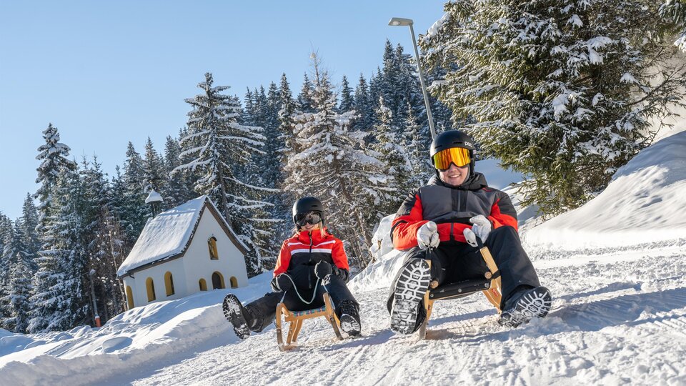Eine verschneite Landschaft mit zwei Personen, die auf Schlitten fahren. Im Hintergrund sind winterliche Bäume und ein kleines, gemütliches Haus zu sehen.