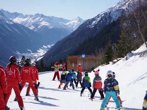 Eine Gruppe von Kindern und Skilehrern auf einer Skipiste in den Bergen. Die Umgebung ist verschneit und es ist sonnig.