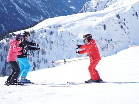Eine Skischule in den Bergen mit drei Personen, die Skifahren lernen. Die Umgebung ist schneebedeckt und die Berge sind beeindruckend.