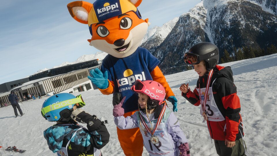 Ein Maskottchen in einem blauen Shirt und einer Mütze steht auf einem schneebedeckten Hang. Um es herum stehen drei Kinder in Skiausrüstung und feiern zusammen.