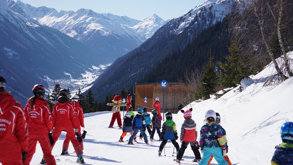 Eine Gruppe von Kindern in bunten Skihosen fährt auf einer verschneiten Piste. Im Hintergrund sind majestätische Berge zu sehen.