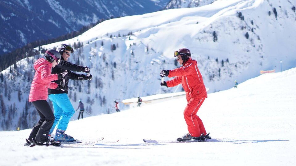 Drei Personen beim Skiunterricht auf einer verschneiten Piste. Im Hintergrund sind beeindruckende Berge zu sehen.