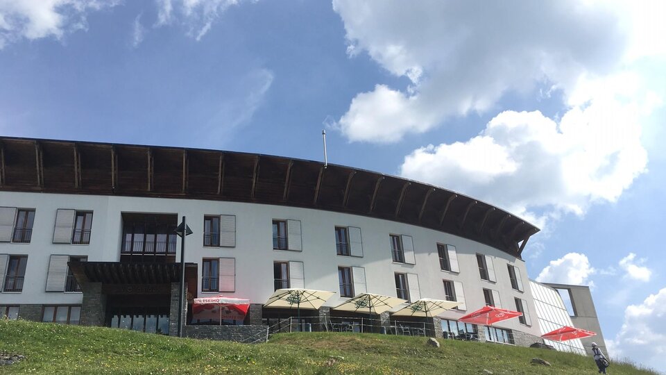 Ein modernes Gebäude in den Bergen, umgeben von Wiesen und blühenden Pflanzen. Der Himmel ist klar und blau.