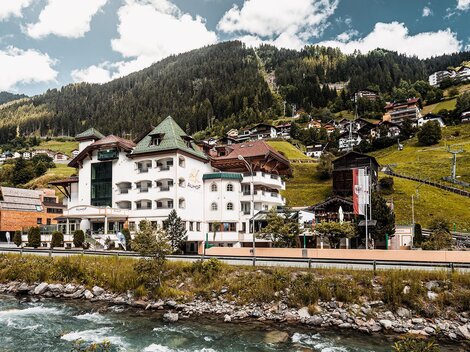 Ein malerisches Hotel am Flussufer, umgeben von grünen Hügeln und Bergen. Die Gebäude sind von schönem, ländlichem Flair geprägt.