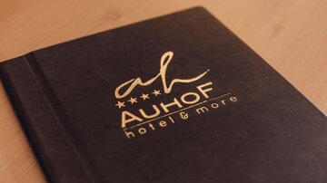 Ein elegantes Notizbuch mit dem Logo des Hotels "Auhof". Es hat eine dunkle Oberfläche und goldene Schriftzüge.