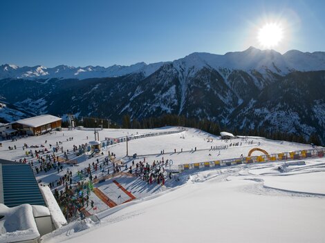 Eine winterliche Berglandschaft mit Skifahrern und sonnigem Himmel. Im Vordergrund sind schneebedeckte Pisten und eine belebte Skischule zu sehen.