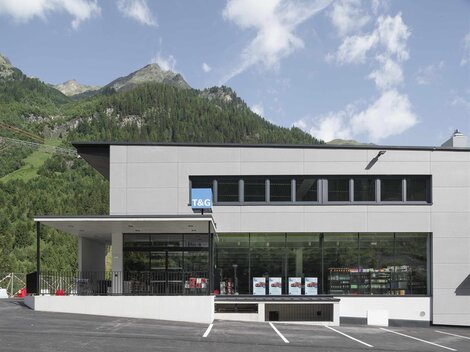 Ein modernes Gebäude mit großen Fenstern und einem Parkplatz davor. Im Hintergrund sind grüne Berge unter einem blauen Himmel zu sehen.