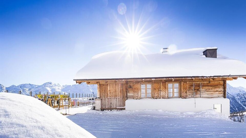 Ein charmantes Holzhaus im Schnee mit strahlender Sonneneinstrahlung. Im Hintergrund sind schneebedeckte Berge sichtbar.