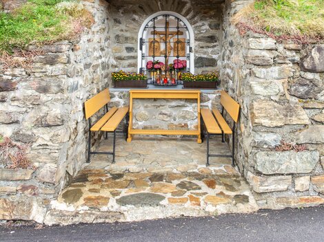 Eine kleine, rustikale Kapelle aus Stein und Holz steht in einer grünen Umgebung. Vor der Kapelle sind zwei Bänke und Blumen zu sehen. | © Bernhard Gruber – Photography