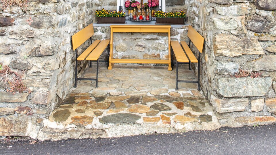 Eine kleine, rustikale Kapelle aus Stein und Holz steht in einer grünen Umgebung. Vor der Kapelle sind zwei Bänke und Blumen zu sehen. | © Bernhard Gruber – Photography
