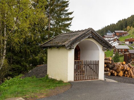 Eine kleine, charmante Hütte mit einem abgewinkelten Dach steht an einem Weg. Im Hintergrund sind Holzstapel und einige Häuser in einer bewaldeten Berglandschaft zu sehen. | © Bernhard Gruber – Photography