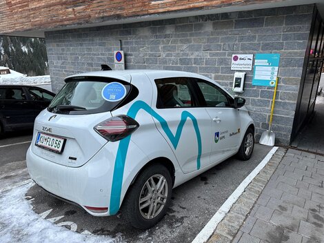 Ein elektrisches Auto parkt an einer Ladestation in einer schneebedeckten Umgebung. Es steht am Rand eines Parkplatzes, umgeben von grauer Steinfassade.